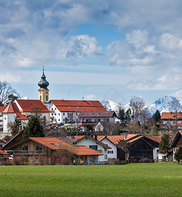 Das Foto zeigt ein bayerisches Dorf mit Kirche und schneebedeckten Bergen im Hintergrund.