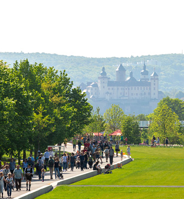 Das Foto zeigt das Gelände der Landesgarten-schau Würzburg mit Schloss im Hintergrund. In der Mitte sieht man eine freie Fläche, die als Kaltluft-schneise frische Luft in die Stadt transportiert. Links unter Baumreihen flanieren viele Menschen. 